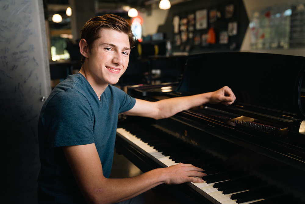 A male student with short, slicked-back brown hair, wearing a dark heather blue T-shirt, sits at a piano.