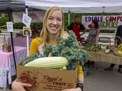 A woman with long blonde hair wearing a white shirt with yellow sleeves holds a box filled with produce at a farmers market. 