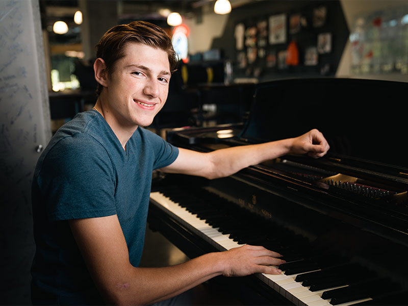 A male student with short, slicked-back brown hair, wearing a dark heather blue T-shirt, sits at a piano.